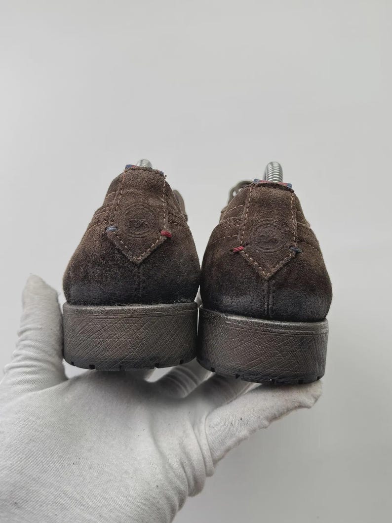 Op de afbeelding: Een paar bruine su&egrave;de schoenen, van achteren bekeken, met een textuurzool. De schoenen hebben een laag profiel en een casual ontwerp. De bovenstukken hebben decoratieve stiksels en kleine accenten van rode en blauwe draad. De schoenen worden vastgehouden door een gehandschoende hand.