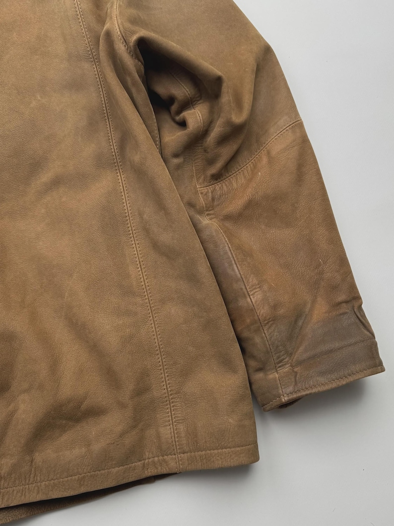 Pu&ograve; includere: Una giacca in pelle color cuoio con una finitura opaca liscia, caratterizzata da cuciture verticali visibili lungo il pannello frontale e una manica lunga con un'area del gomito rinforzata. Il capo presenta un colore marrone chiaro uniforme.