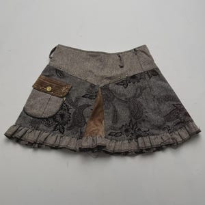 Op de afbeelding: Een bruine en grijze minirok met een donker bloemrijk paisley-patroon, een klein klepzakje met een goudkleurige knoop, een bruin suède-achtig driehoekig inzetstuk, een geplooide zoom en een brede tailleband met kleine metalen studs.