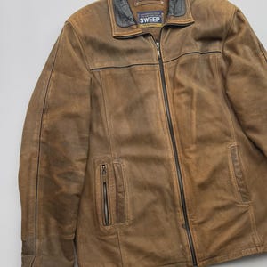 Pu&ograve; includere: Una giacca in pelle marrone con colletto nero, chiusura frontale con zip integrale e due tasche verticali con zip sul davanti. L'etichetta interna riporta il testo SWEEP Young Fashion. La giacca presenta cuciture e pannelli visibili.
