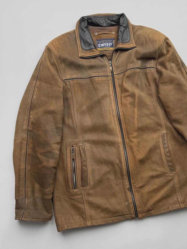 Pu&ograve; includere: Una giacca in pelle marrone con colletto nero, chiusura frontale con zip integrale e due tasche verticali con zip sul davanti. L'etichetta interna riporta il testo SWEEP Young Fashion. La giacca presenta cuciture e pannelli visibili.