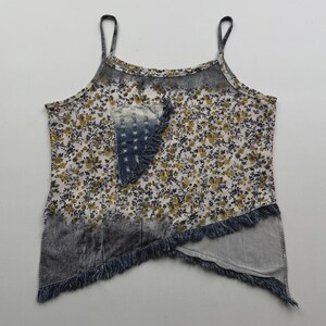 Può includere: Un top smanicato con base bianca e una piccola stampa floreale gialla e nera. Il capo presenta una toppa in denim blu scuro consumato sul petto e un orlo diagonale rifinito con frange in denim sfilacciate lungo il bordo inferiore.