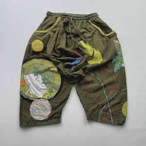 May include: Olive green shorts with a drawstring waist and pockets. The shorts feature colorful embroidered patches and appliqués, including a circular design with a whimsical illustration and floral accents. The shorts have a relaxed fit.