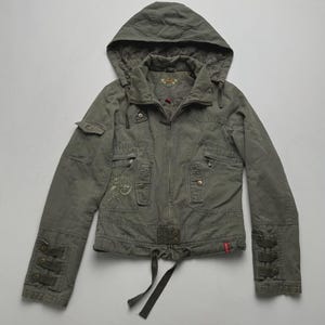 Peut inclure: Une veste à capuche vert olive dotée d'une fermeture éclair intégrale, de multiples poches utilitaires et d'un ourlet à cordon de serrage. Les manches sont ornées de trois brides décoratives et boutons aux poignets, avec une broderie florale sur la poitrine.