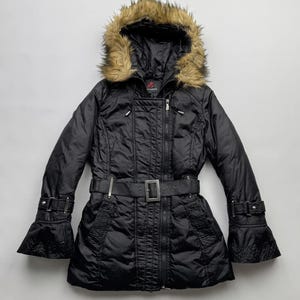 Könnte beinhalten: Schwarze, gegurtete Steppjacke mit einer Kapuze, die mit Kunstfell gefüttert ist. Die Jacke hat einen Reißverschluss und einen Gürtel mit silberner Schnalle.