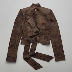 May include: A brown long-sleeved jacket featuring a high collar and a tie-front closure. The garment is covered in a dark brown tonal graphic print consisting of repeated text and abstract patterns across the entire surface of the fabric.