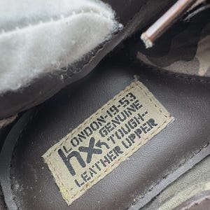 Op de afbeelding: Close-up van de binnenkant van een bruine leren schoen. Een label leest "LONDON-19-53 GENUINE TOUGH LEATHER UPPER" met het "hx" logo. De schoen heeft een witte voering en een camouflagepatroon.