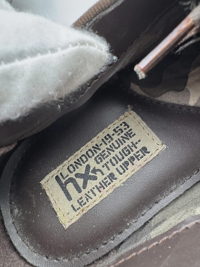 Op de afbeelding: Close-up van de binnenkant van een bruine leren schoen. Een label leest "LONDON-19-53 GENUINE TOUGH LEATHER UPPER" met het "hx" logo. De schoen heeft een witte voering en een camouflagepatroon.