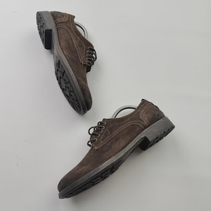 Op de afbeelding: Twee bruine su&egrave;de herenschoenen met donkergrijze zolen en veters worden getoond tegen een witte achtergrond. De schoenen hebben een klassiek ontwerp met een textuur. De zolen hebben een robuust profiel.