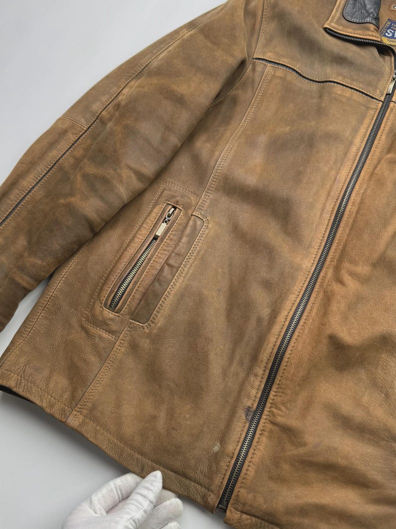 Pu&ograve; includere: Una giacca in pelle color cuoio con chiusura frontale a zip, una tasca verticale con zip sul petto e un colletto marrone scuro. La giacca presenta cuciture visibili lungo i pannelli e una finitura testurizzata e vissuta sulla pelle.