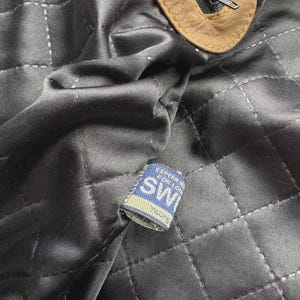 Pu&ograve; includere: Una fodera in tessuto nero trapuntato con un tirazip in pelle marrone chiaro e una piccola etichetta tessuta blu e bianca con la scritta "SW" e "Young", con ulteriore testo parzialmente oscurato dalla piega del tessuto.