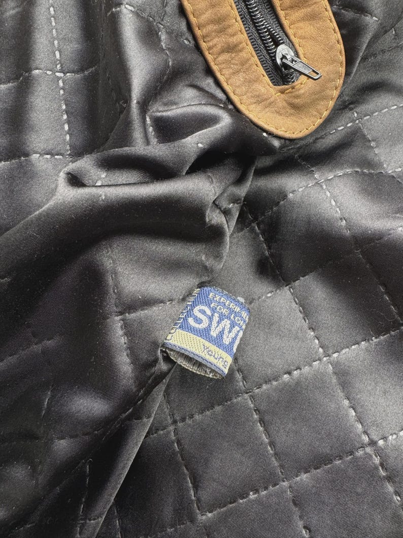Pu&ograve; includere: Una fodera in tessuto nero trapuntato con un tirazip in pelle marrone chiaro e una piccola etichetta tessuta blu e bianca con la scritta "SW" e "Young", con ulteriore testo parzialmente oscurato dalla piega del tessuto.