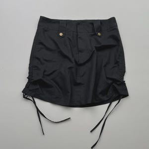May include: A black mini skirt with two gold-colored buttons at the waistband. The skirt has drawstring details on each side, adding a unique touch to the design. The fabric appears to be a lightweight material.