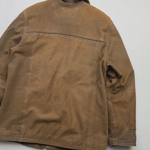 Pu&ograve; includere: Una giacca in camoscio marrone con colletto ripiegato, maniche lunghe e una cucitura orizzontale sulla parte superiore della schiena. Il capo presenta dettagli di cuciture visibili lungo le spalle e i pannelli posteriori, con una texture opaca.