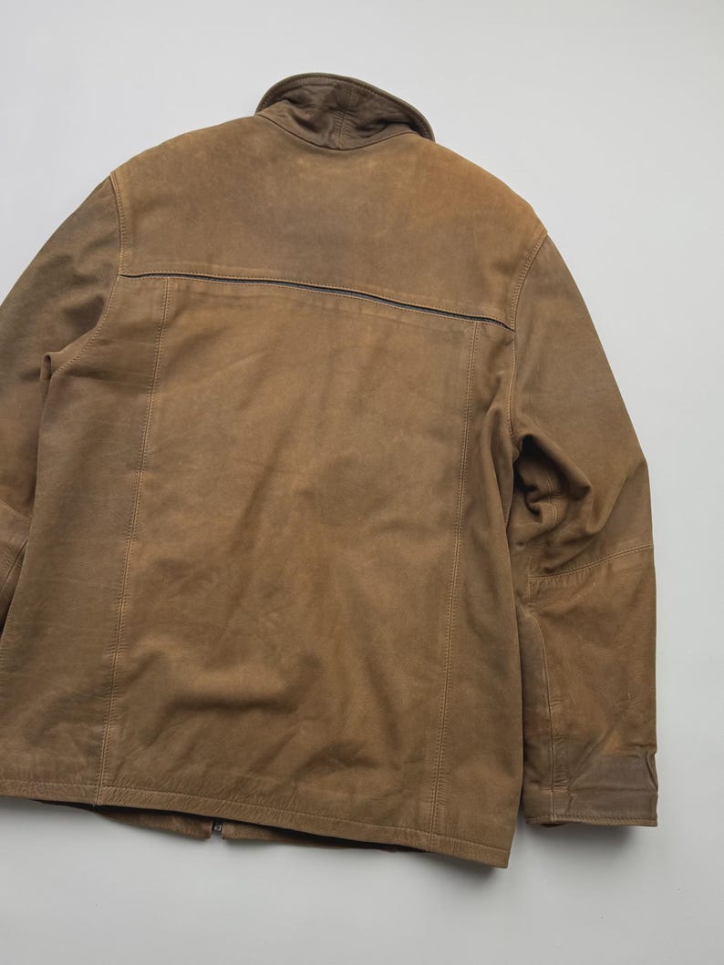 Pu&ograve; includere: Una giacca in camoscio marrone con colletto ripiegato, maniche lunghe e una cucitura orizzontale sulla parte superiore della schiena. Il capo presenta dettagli di cuciture visibili lungo le spalle e i pannelli posteriori, con una texture opaca.