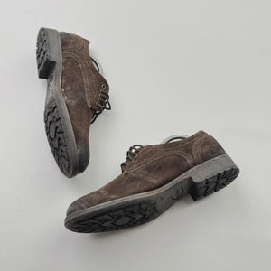 Op de afbeelding: Twee bruine su&egrave;de leren schoenen met donkergrijze zolen en veters. De schoenen hebben een robuuste, getextureerde uitstraling en zijn ontworpen voor dagelijks gebruik. De zolen hebben een patroonprofiel voor grip.