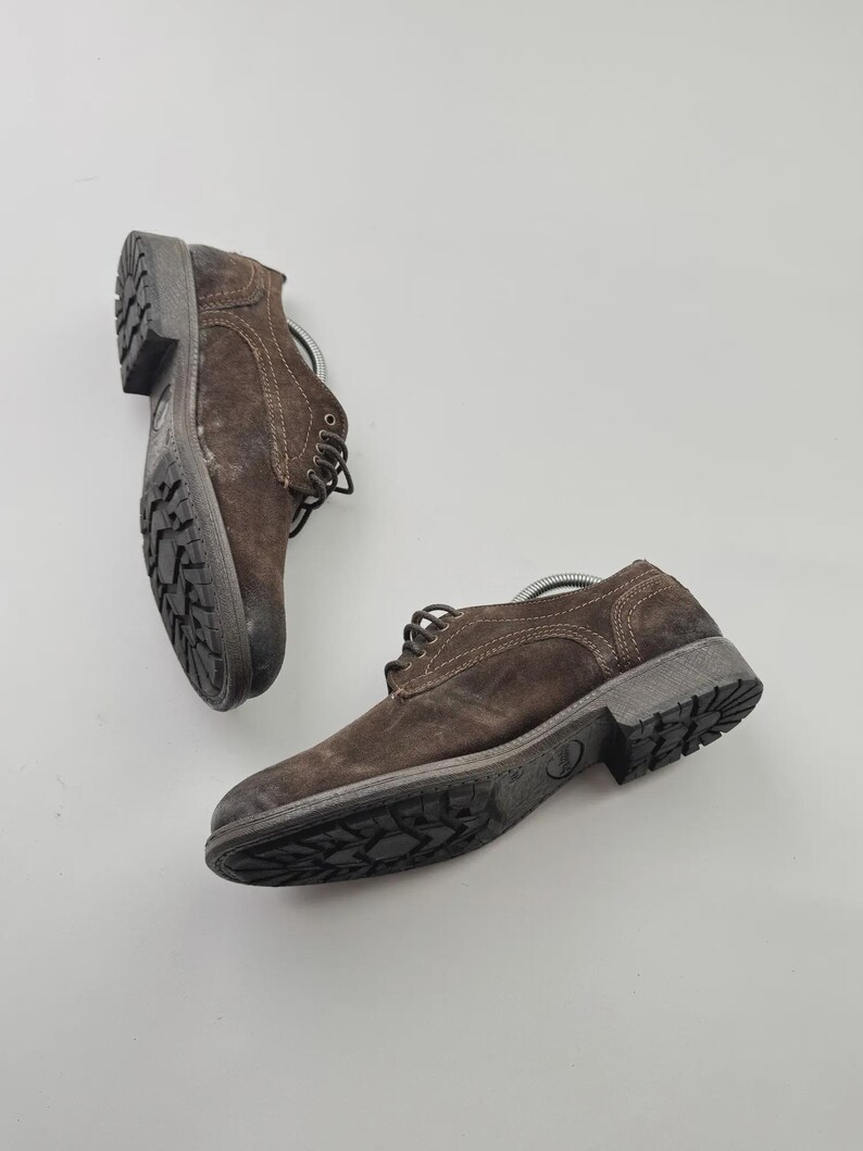Op de afbeelding: Twee bruine su&egrave;de leren schoenen met donkergrijze zolen en veters. De schoenen hebben een robuuste, getextureerde uitstraling en zijn ontworpen voor dagelijks gebruik. De zolen hebben een patroonprofiel voor grip.