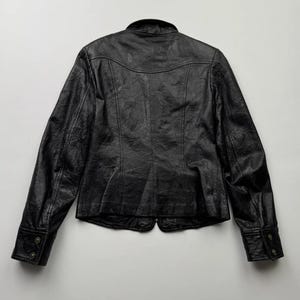 以下が含まれることがあります： 襟付き、長袖、ボタン留めの黒い革のジャケット。ジャケットは、やや体にフィットしたシルエットで、表面にテクスチャがあります。