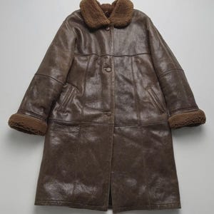 Könnte beinhalten: Ein langer, brauner Ledermantel mit einem Kragen und Manschetten aus Lammfell. Der Mantel hat eine Knopfleiste und zwei Taschen. Der Mantel ist im Vintage-Stil gehalten.
