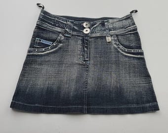 Puede incluir: Una falda vaquera de lavado oscuro con un diseño clásico de cinco bolsillos. La falda presenta cierre de botón y cremallera, trabillas para el cinturón y detalles de pedrería decorativos en los bolsillos y la cintura. La falda tiene un aspecto ligeramente descolorido.