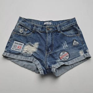 Puede incluir: Pantalones cortos vaqueros azules con detalles desgastados, incluyendo una sección rasgada y parches. Los pantalones cortos tienen cierre de botón, bolsillos y dobladillos. Los parches incluyen texto como "HAOER 33" y "UNION 1987 MADE".