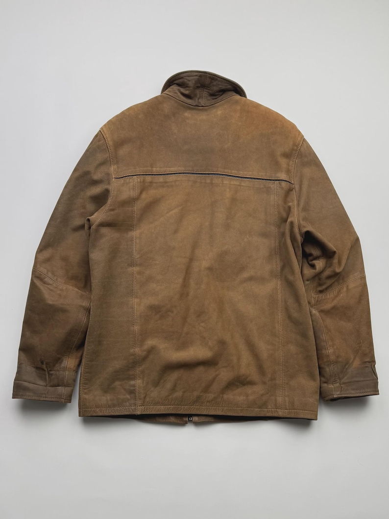 Pu&ograve; includere: Una giacca in camoscio marrone con colletto alto, maniche lunghe con polsini e una cucitura orizzontale sulla parte superiore della schiena. Il capo presenta dettagli con cuciture a vista lungo i pannelli verticali e una texture liscia e opaca.