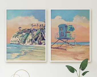May include: Two framed wall art prints featuring coastal scenes. The left print depicts a cliffside with palm trees and ocean waves. The right print shows a blue lifeguard tower labeled 20 on a sandy beach under a soft pink and blue sky.