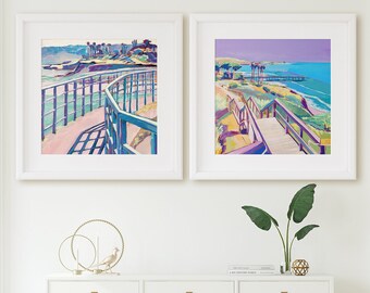 May include: Two framed abstract paintings of a coastal scene with a boardwalk and ocean view. The paintings are hung on a white wall above a white console table with gold legs. A green velvet chair is visible in the doorway on the right.