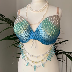 May include: A mermaid-style bra with a scale pattern in blue and green hues, presented on a mannequin. It features a crystal pendant and layered necklaces of pearls and crystals. This bra is a unique piece of wearable art.