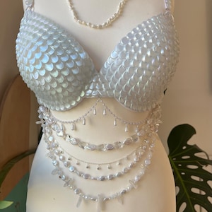 Puede incluir: Un conjunto de sujetador y cadena de cintura blanco nacarado, exhibido en un maniquí. El sujetador presenta un patrón similar a escamas. La cadena de cintura está adornada con perlas y detalles de cristal transparente. También se incluye un collar de perlas en capas.