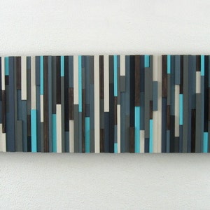 Pu&ograve; includere: Arte da parete astratto realizzato in legno con una variet&agrave; di tonalit&agrave; di blu, grigio, marrone e bianco. Il legno &egrave; disposto in strisce verticali per creare un design moderno e geometrico.