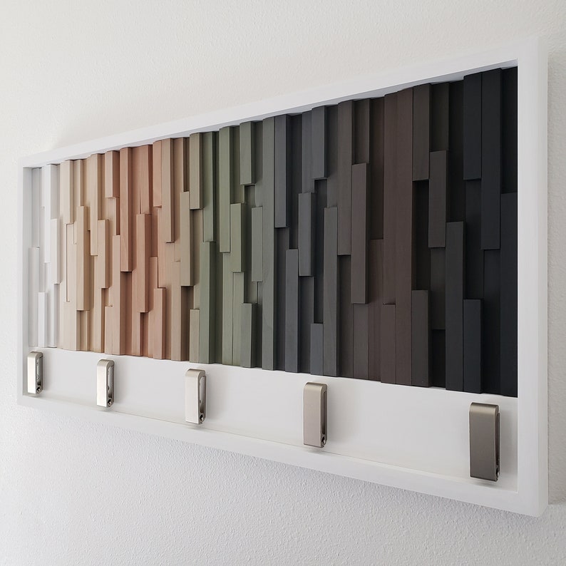 May include: A white framed wall mounted coat rack with a geometric wood design in shades of beige, green, gray, and black. The rack has five silver hooks.