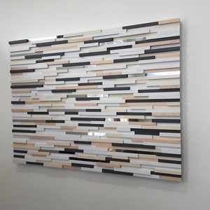May include: A wall art piece made of wood with a variety of colors and textures. The piece is made up of many small rectangular pieces of wood, arranged in a staggered pattern. The colors of the wood include black, white, brown, and beige.