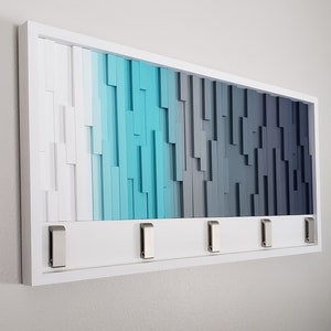 May include: White wood wall art with a gradient of blue and gray colors. The art has five metal hooks on the bottom.