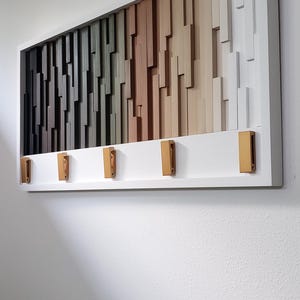 May include: A white framed wall art piece with a geometric design of wooden blocks in shades of gray, green, brown, and beige. The piece has five gold hooks on the bottom.