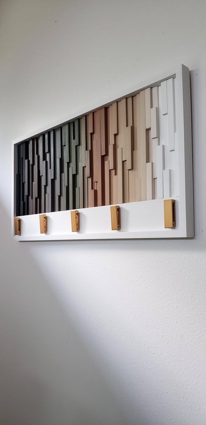 May include: A white framed wall art piece with a geometric design of wooden blocks in shades of gray, green, brown, and beige. The piece has five gold hooks on the bottom.