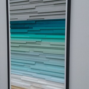 May include: A framed wood wall art piece with a layered design. The piece features a gradient of colors from gray to teal to white to tan, resembling a beach scene.