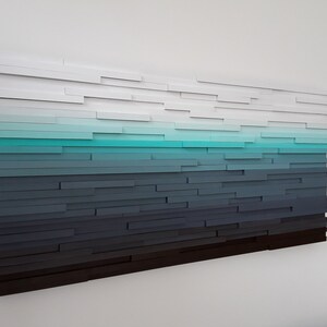 May include: Abstract wall art made of wood with a gradient of colors from white to black, with a turquoise blue band in the middle.