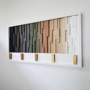 May include: A white framed wall art piece with a gradient of wood tones. The wood is arranged in a staggered pattern, creating a modern and abstract design. The piece has five gold hooks on the bottom.