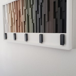 May include: A framed wall art piece with a wood mosaic design. The mosaic features a gradient of colors from white to black, with shades of brown, beige, green, and gray. The frame is white and has five black hooks on the bottom.