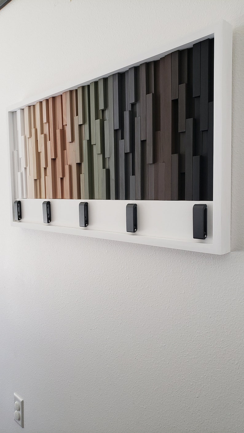 May include: A framed wall art piece with a wood mosaic design. The mosaic features a gradient of colors from white to black, with shades of brown, beige, green, and gray. The frame is white and has five black hooks on the bottom.