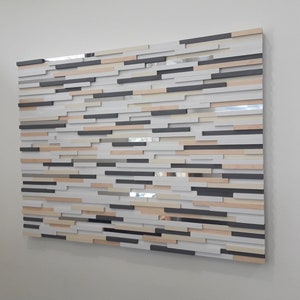 May include: A wall art piece made of wood with a variety of colors and textures. The piece is made up of many small, rectangular pieces of wood that are arranged in a random pattern. The colors of the wood range from light beige to dark brown. The piece is mounted on a white wall.