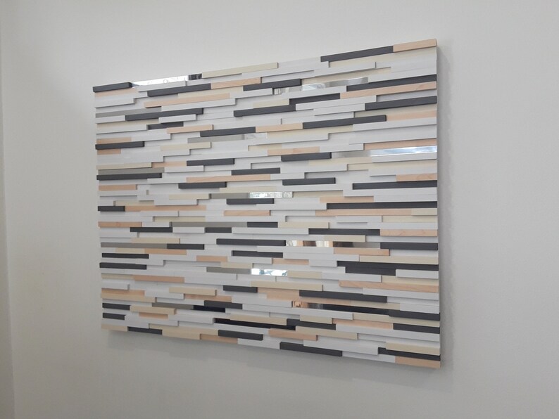 May include: A wall art piece made of wood with a variety of colors and textures. The piece is made up of many small, rectangular pieces of wood that are arranged in a random pattern. The colors of the wood range from light beige to dark brown. The piece is mounted on a white wall.