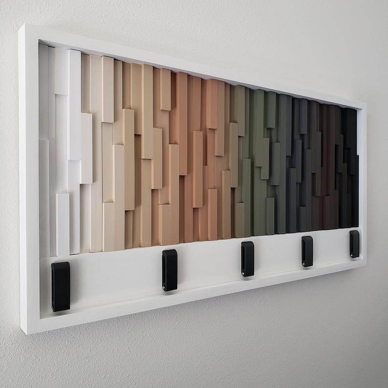 May include: A white wooden wall mounted key holder with a geometric design in shades of white, beige, brown, green, gray, and black. Five black hooks are attached to the bottom of the holder.