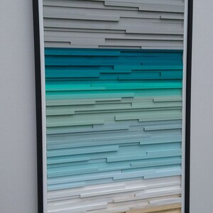 May include: A framed abstract art piece with a gradient of colors, starting with gray at the top, transitioning to teal, then light blue, and ending with a beige color at the bottom. The piece is made of wood slats.