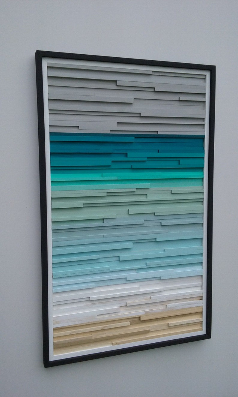 May include: A framed abstract art piece with a gradient of colors, starting with gray at the top, transitioning to teal, then light blue, and ending with a beige color at the bottom. The piece is made of wood slats.