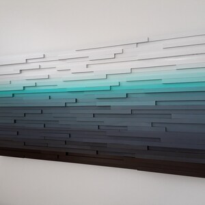 May include: Abstract wall art made of wood with a gradient of colors from white to dark gray, with a turquoise blue band in the middle.