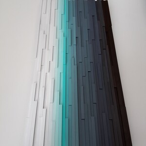 May include: Abstract wall art made of wood painted in shades of white, turquoise, blue, gray, and black. The wood pieces are arranged in vertical rows to create a gradient effect.
