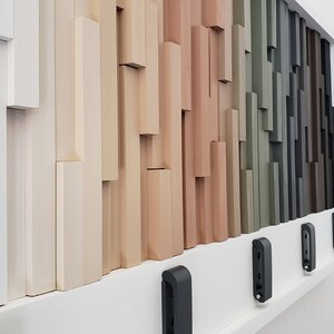 May include: A wall display of various wood samples in different shades of white, beige, brown, green, and gray. The samples are arranged in a staggered pattern, creating a visual texture. There are four black doorbells mounted on the wall below the wood samples.