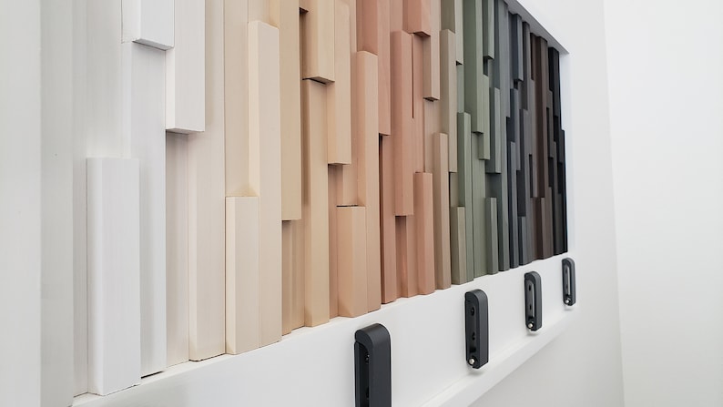 May include: A wall display of various wood samples in different shades of white, beige, brown, green, and gray. The samples are arranged in a staggered pattern, creating a visual texture. There are four black doorbells mounted on the wall below the wood samples.